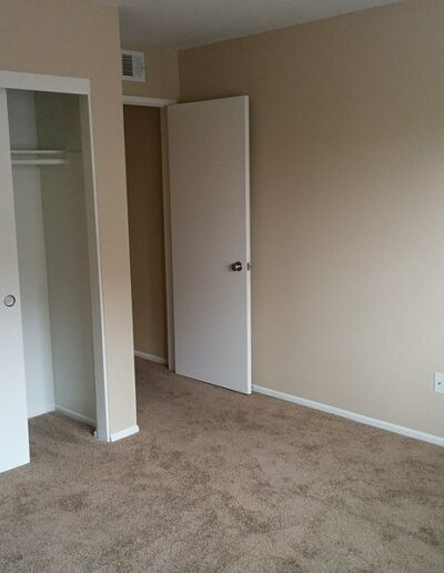 Empty room with beige walls, tan carpet, an open white closet with sliding doors, and a slightly open white door leading to another space. Electrical outlets and a smoke detector are visible on the walls.