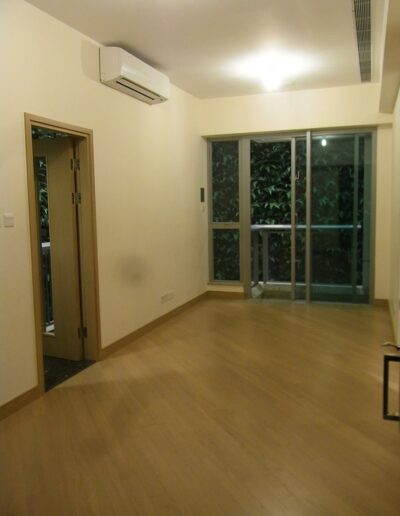 A small, empty room with light wood flooring, cream-colored walls, a ceiling light, wall-mounted air conditioner, open door to the left, and a glass sliding door leading to a balcony with plants outside.