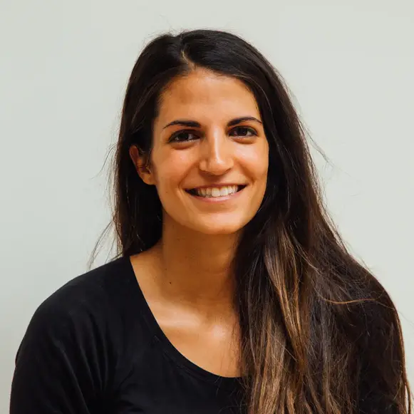 A woman with long, dark hair and a black top smiles at the camera against a plain, light background.