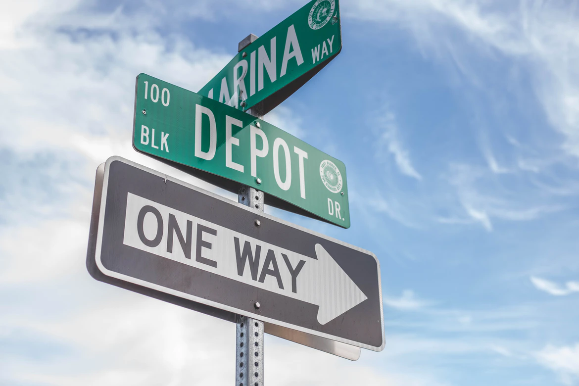 Close-up of street signs at an intersection, showing Depot Dr, Marina Way, and a One Way sign with an arrow pointing right, against a partly cloudy blue sky.