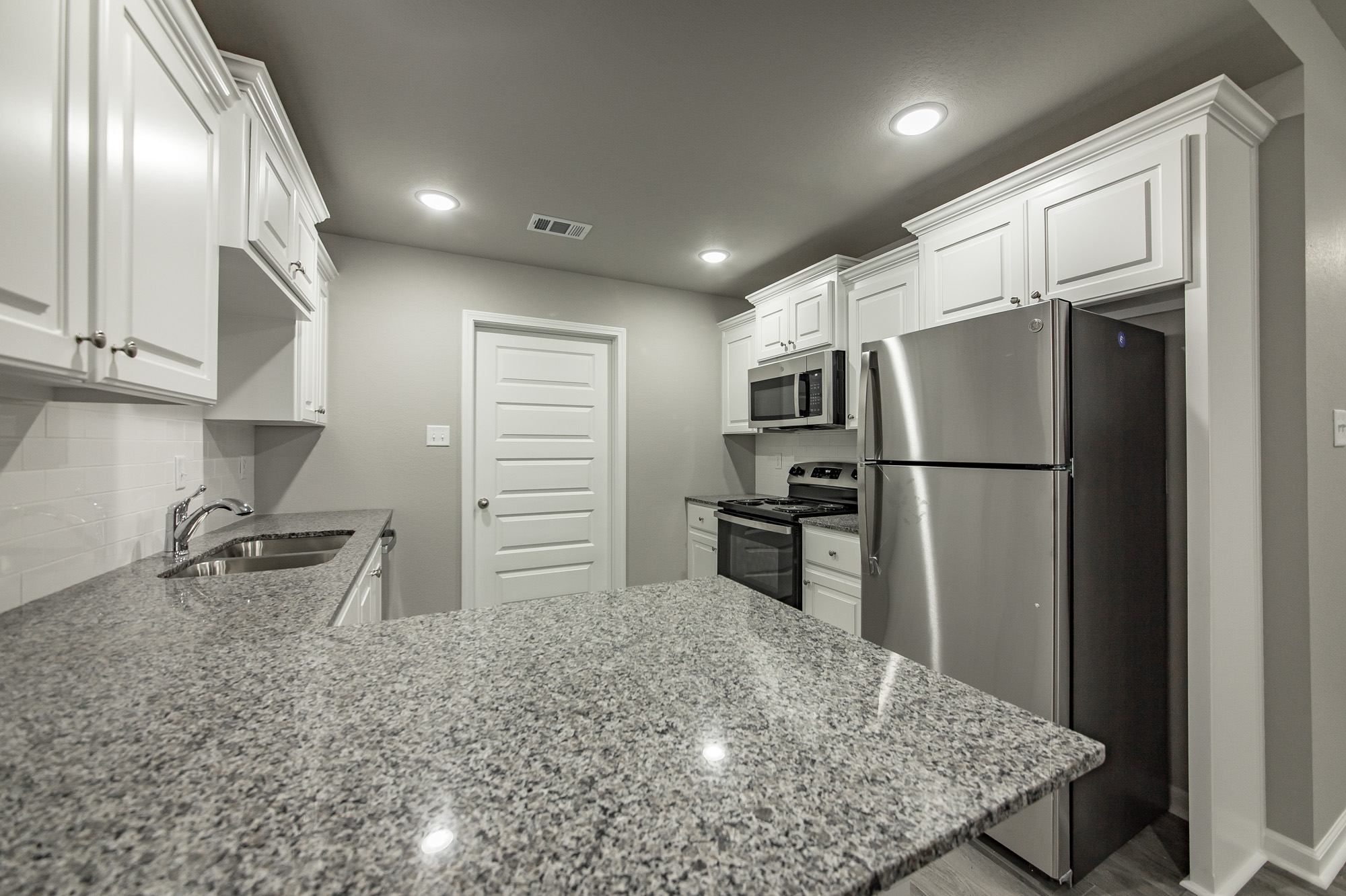 Modern kitchen with white cabinets, stainless steel appliances including a refrigerator, oven, and microwave, granite countertops, under-cabinet sink, and neutral gray walls with recessed lighting.
