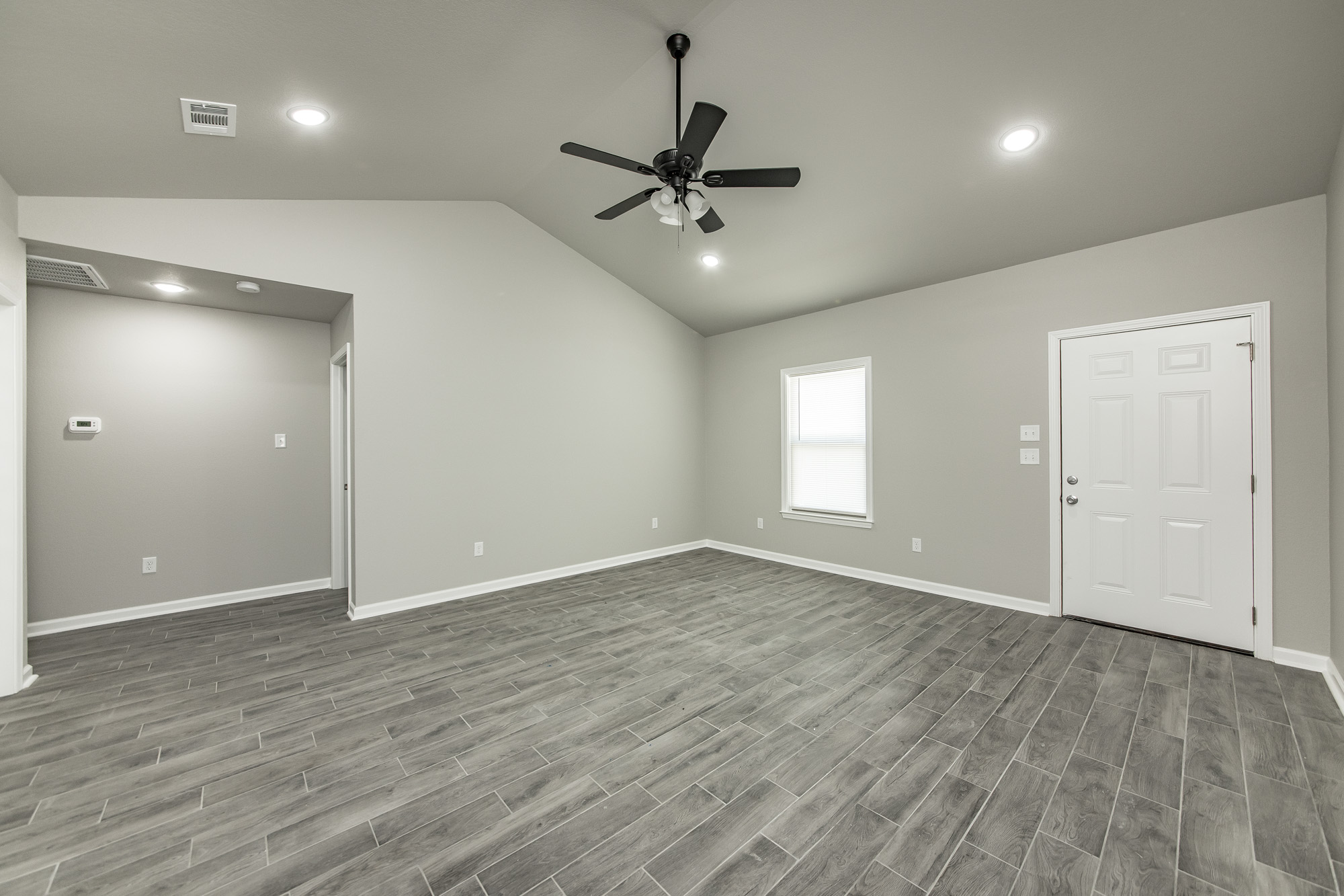 A modern, empty living room with gray walls and wood-look tile flooring, featuring a white door, a window, recessed lights, and a black ceiling fan. The space has a vaulted ceiling and neutral decor.