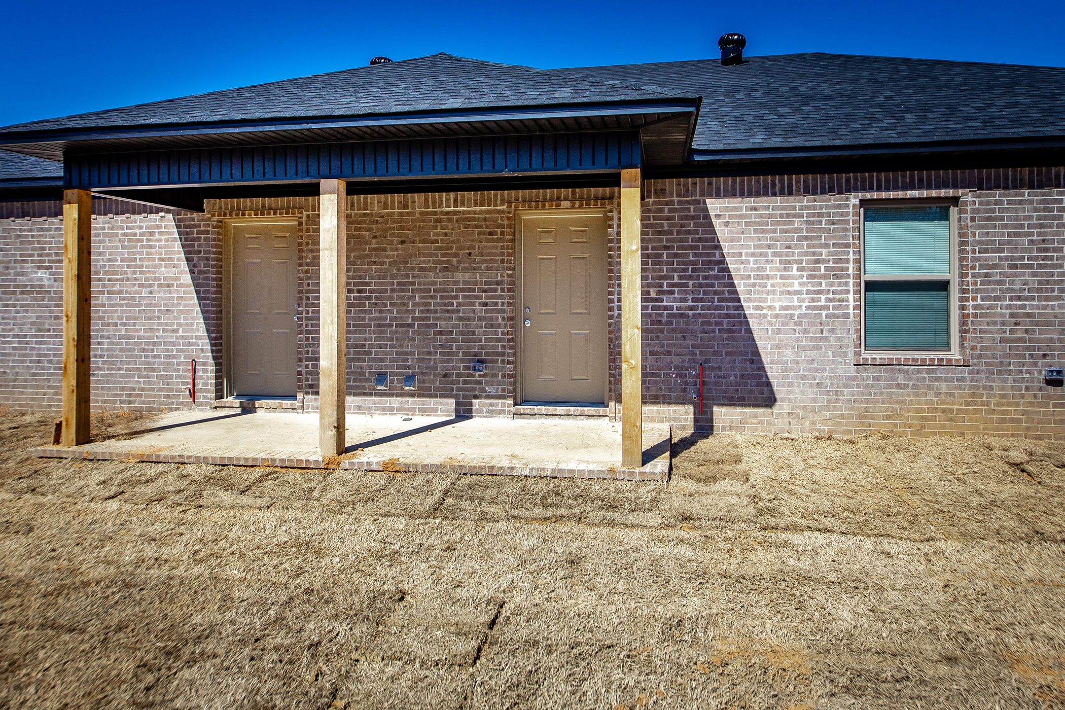 A newly constructed brick house with two beige doors, a small covered porch supported by wooden posts, a single window, and dry, freshly-laid sod in the yard under a clear blue sky.