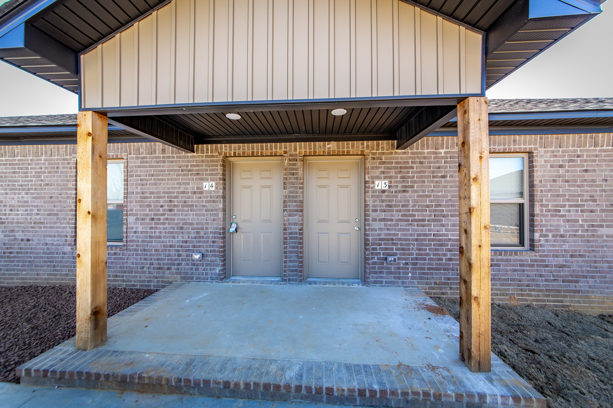 Two tan doors with silver handles in a brick duplex, marked 04 and 05, under a covered entry supported by wooden posts. There are small windows on each side and a concrete porch.