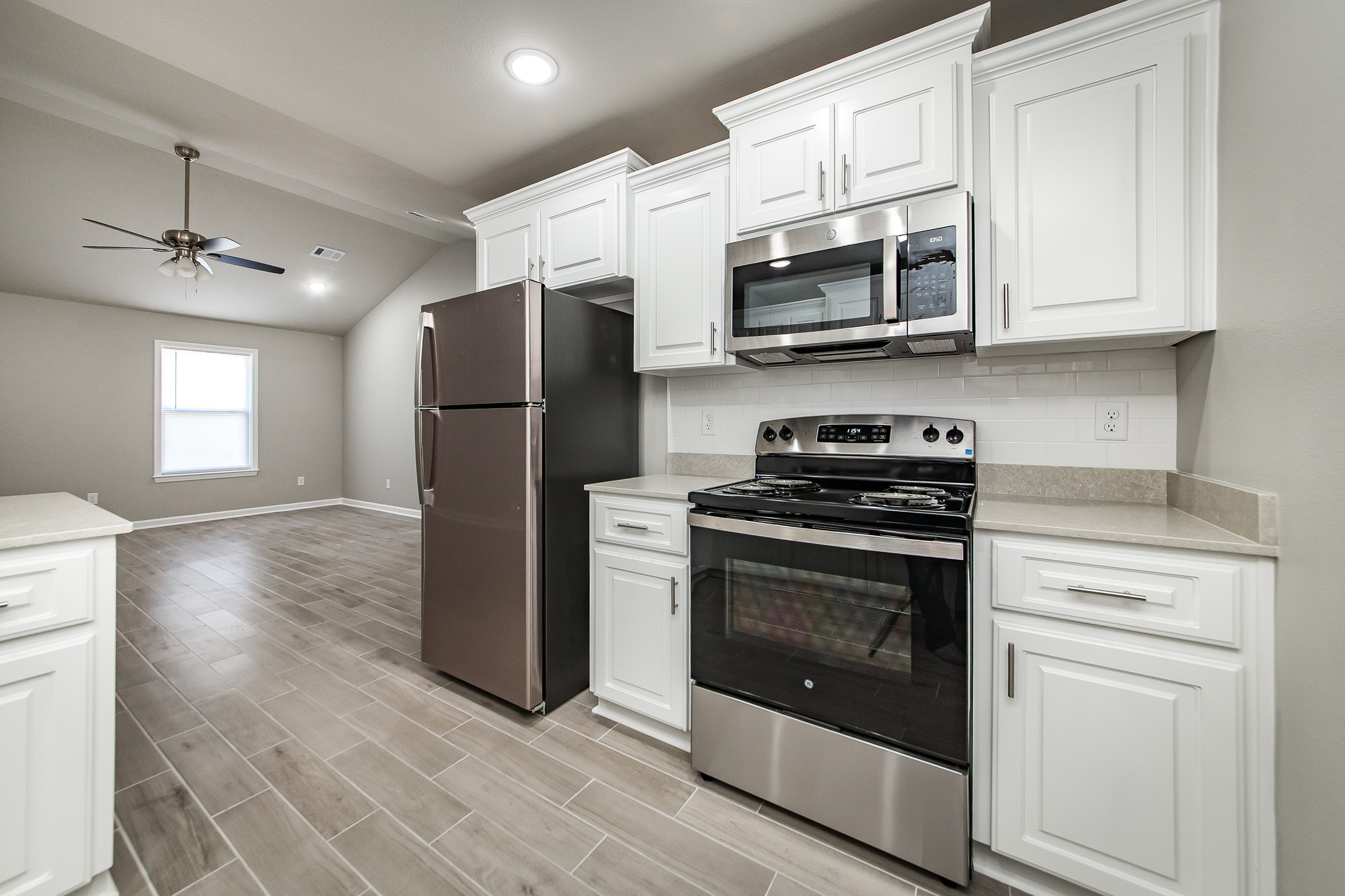 Modern kitchen with white cabinets, stainless steel appliances including a refrigerator, stove, and microwave, light beige countertops, and light wood-look tile flooring; open to a living area with a ceiling fan.