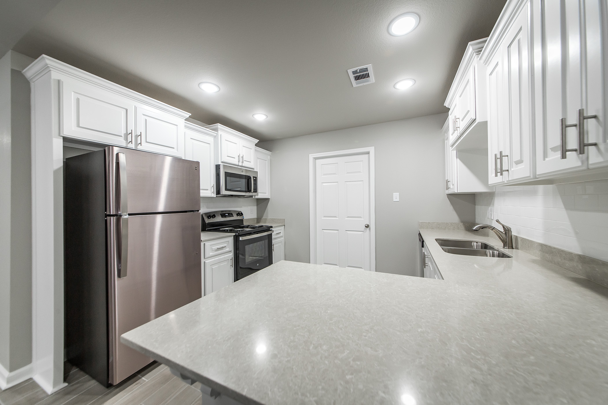 Modern kitchen with white cabinets, stainless steel appliances including a refrigerator, oven, and microwave, light gray countertops, under-cabinet lighting, and a central island under recessed ceiling lights.