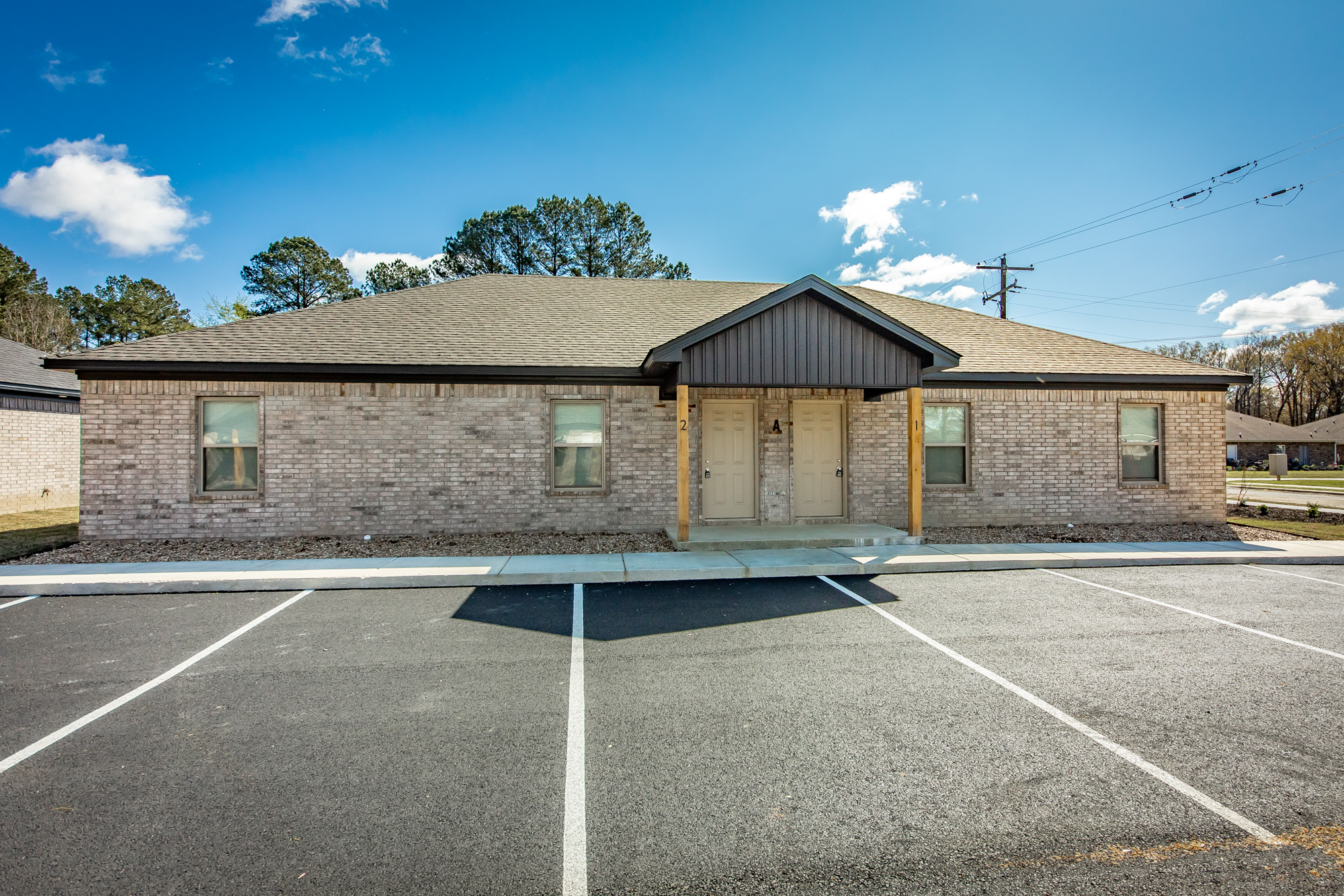 Aerial view of a small new apartment complex with several single-story buildings, freshly paved parking lots, landscaped areas, and a few parked cars, surrounded by grass and trees.