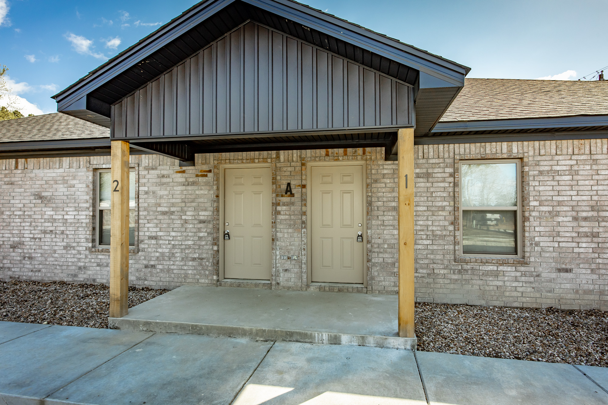 A duplex building with two beige doors labeled 1 and A, separated by wooden posts under a shared covered entryway, brick exterior walls, and a concrete porch.