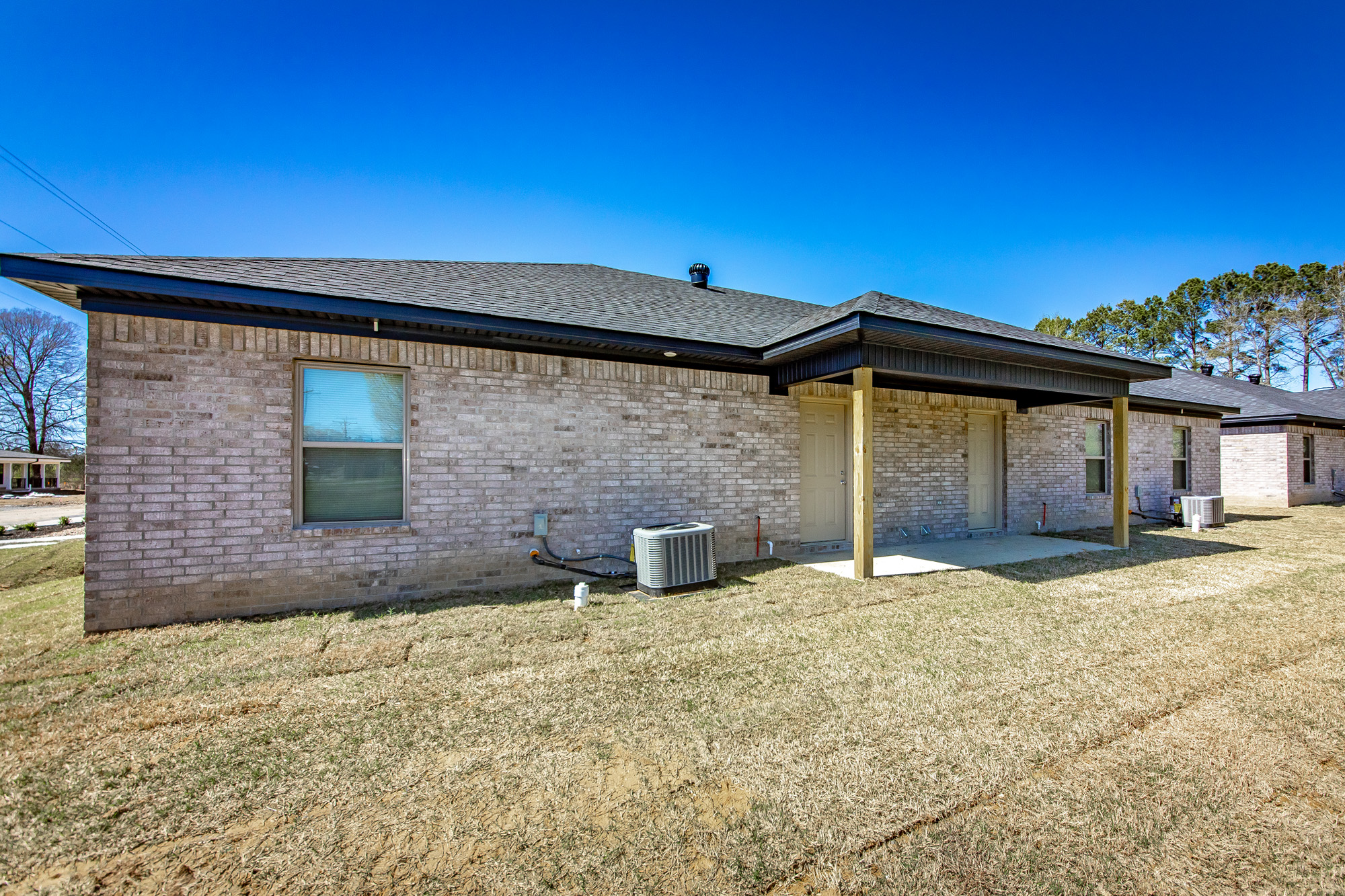 A single-story brick house with a black roof, two windows, an air conditioning unit, and a small covered patio in the backyard. The lawn is dry and patchy under a clear blue sky.