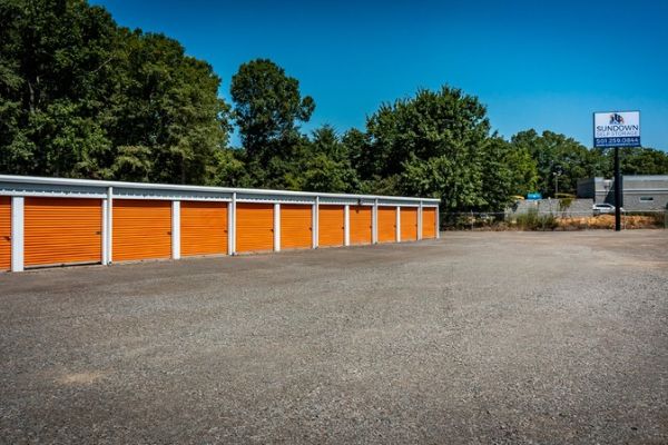 A row of orange self-storage units with white roll-up doors in a gravel lot under a clear blue sky, with a Sundown Self Storage sign in the background.