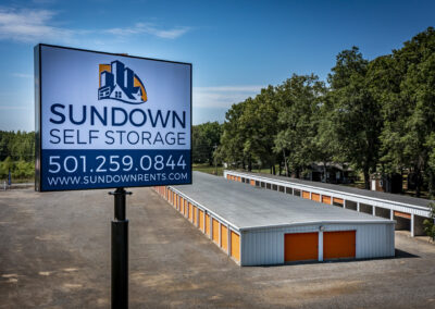 A sign for Sundown Self Storage featuring the business name, phone number, and website, overlooking a row of storage units.