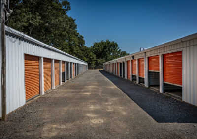 A gravel aisle between two rows of metal self-storage units with orange roll-up doors.